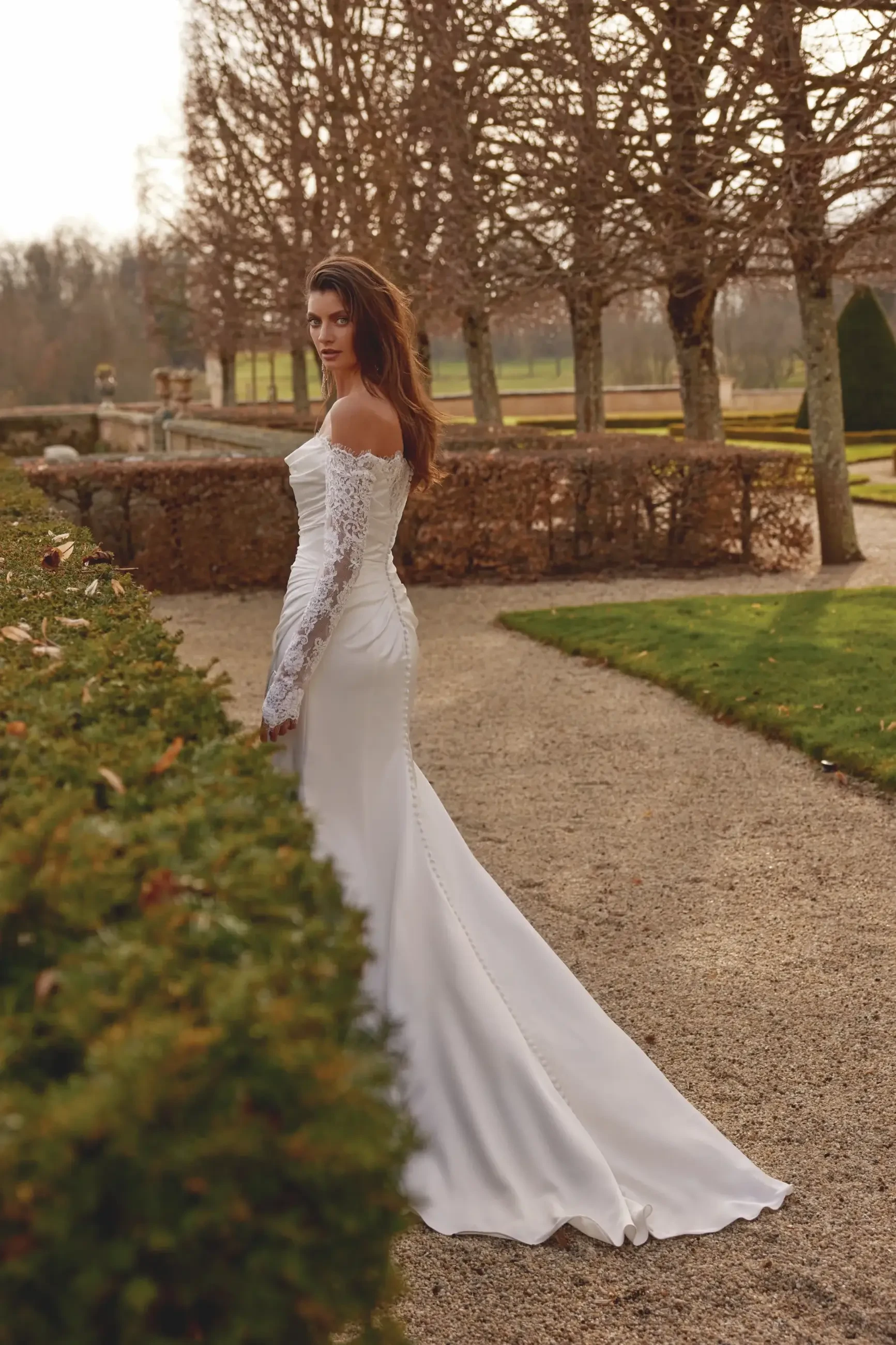 Model is wearing a long-sleeve charmeuse fit-and-flare wedding gown with an off-the-shoulder neckline.