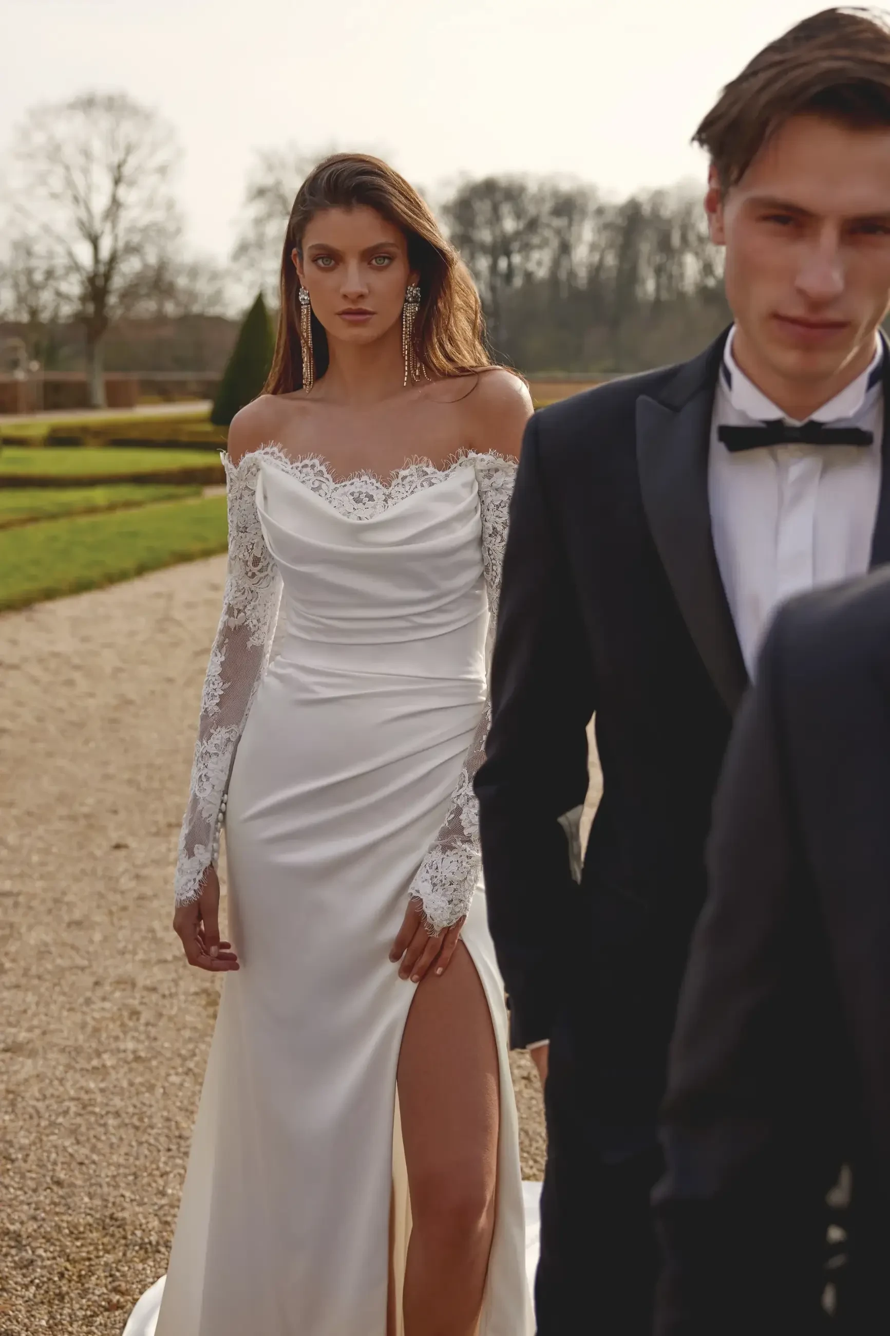 Model is wearing a long-sleeve charmeuse fit-and-flare wedding gown with an off-the-shoulder neckline.