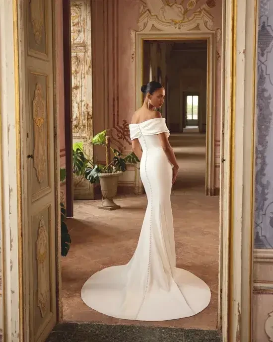 Model is wearing a charmeuse fit-and-flare wedding gown with a pleated portrait neckline.