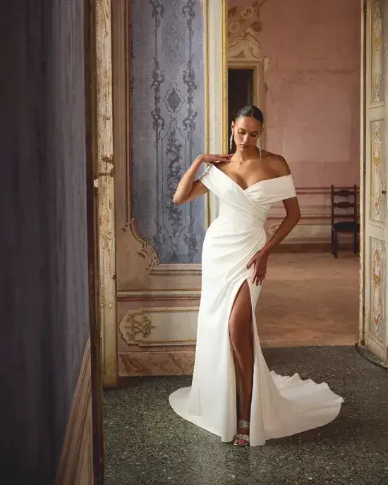 Model is wearing a charmeuse fit-and-flare wedding gown with a pleated portrait neckline.