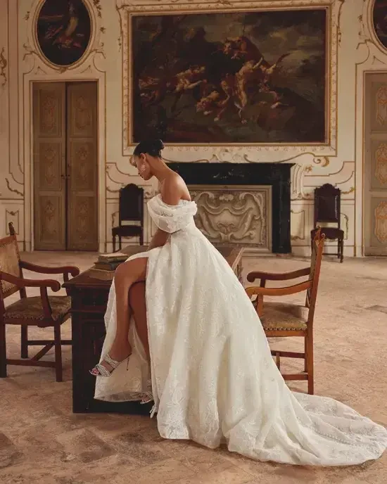 Model is wearing an embroidered crinkled organza A-line wedding gown with pockets and a detachable off-the-shoulder sleeve.
