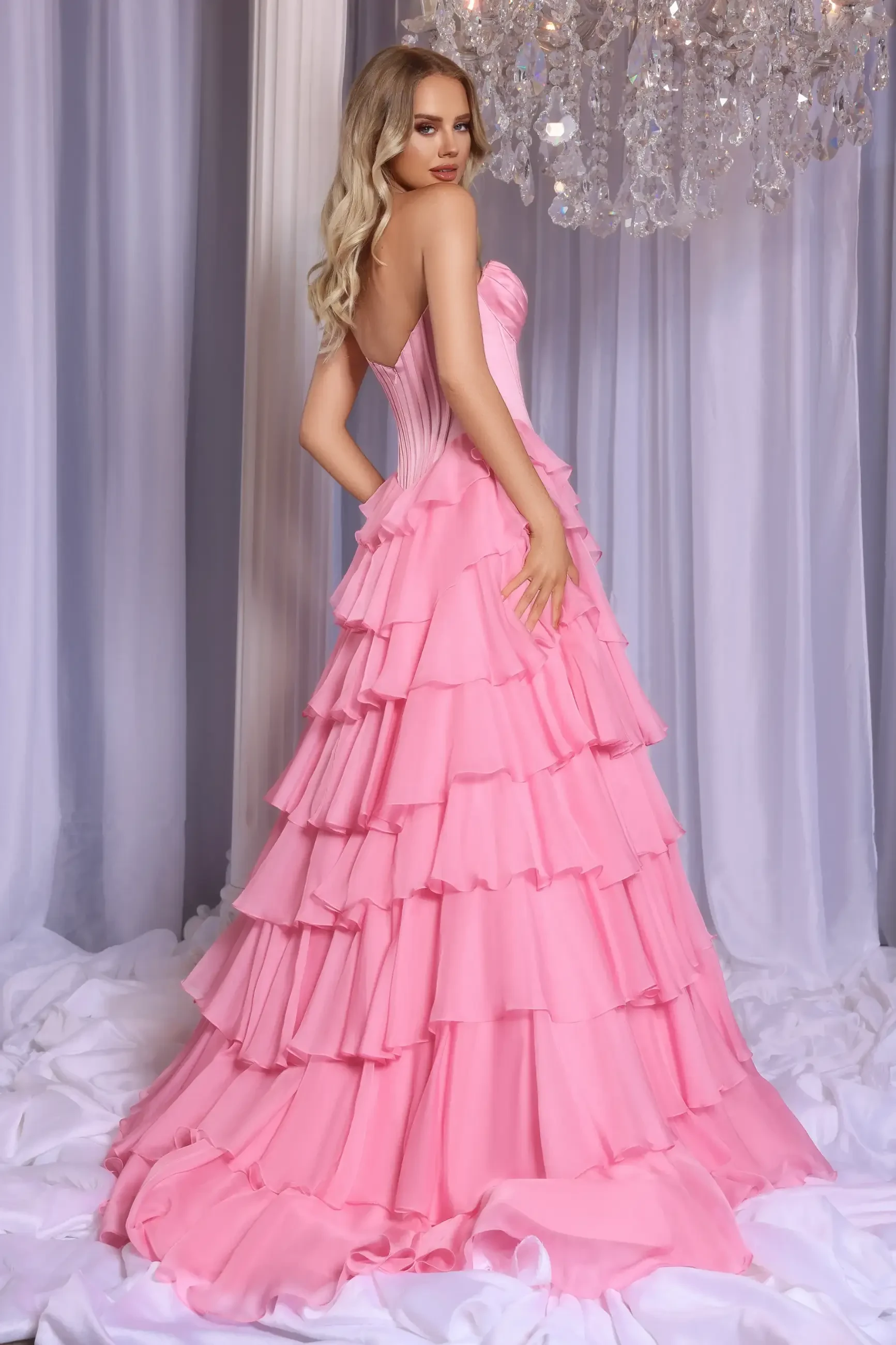 Woman in a glamorous, tiered pink gown poses under a sparkling chandelier. The elegant setting includes soft, flowing drapery, creating a luxurious atmosphere.