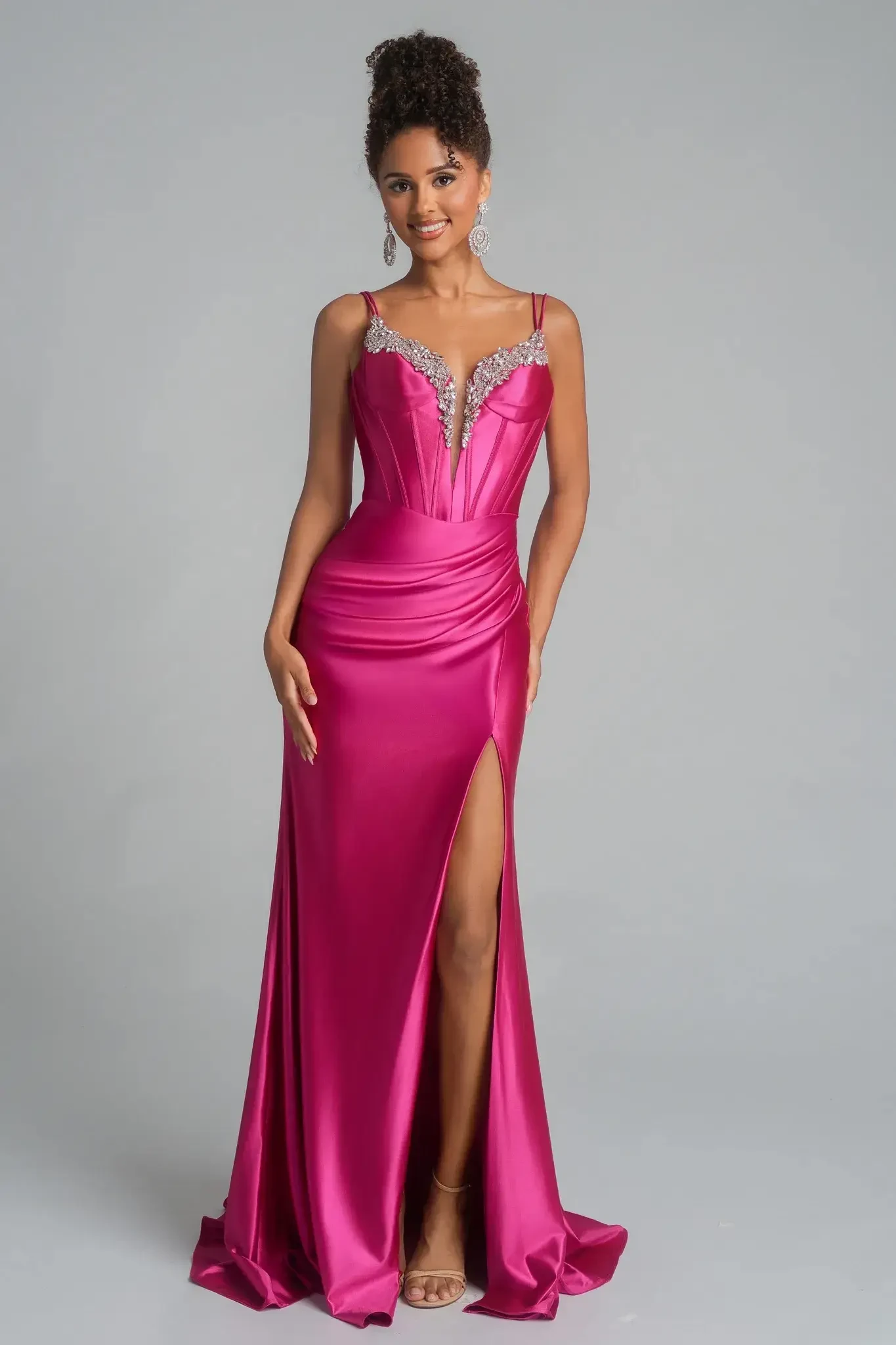 A woman wears a vibrant pink satin gown with a high slit and jeweled neckline. She looks elegant, exuding confidence, set against a neutral gray background.